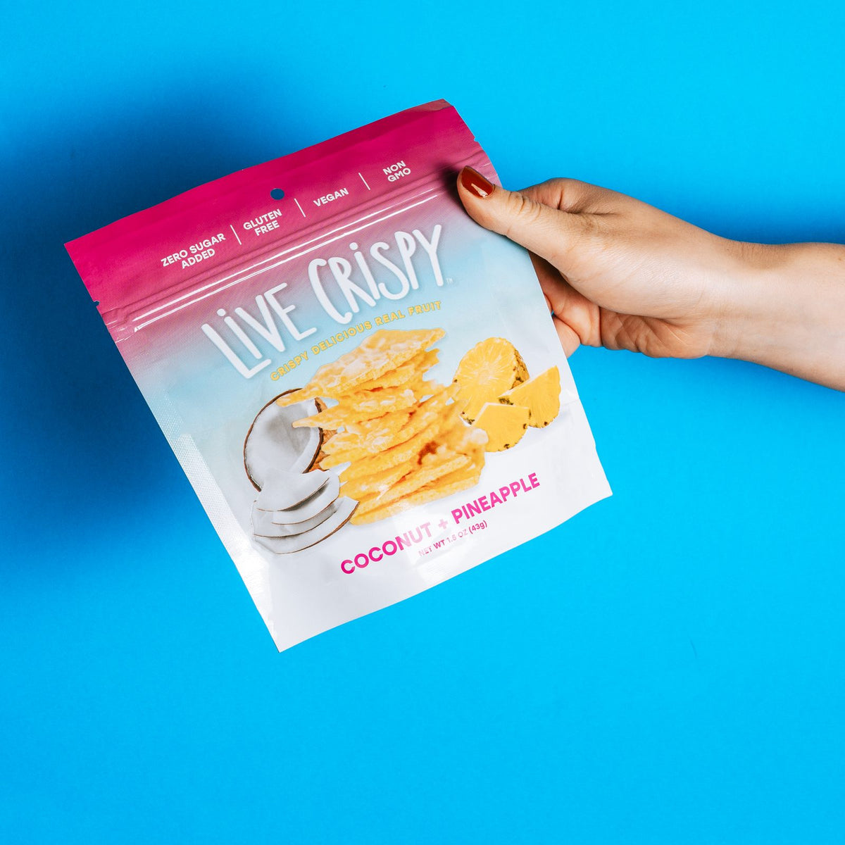 A hand holding a bag of Live Crispy Coconut and Pineapple against a blue background